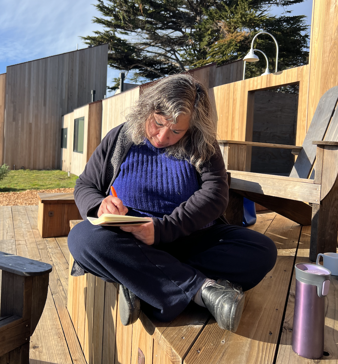 A woman writing in a notebook on a wooden deck, cypress trees in the background