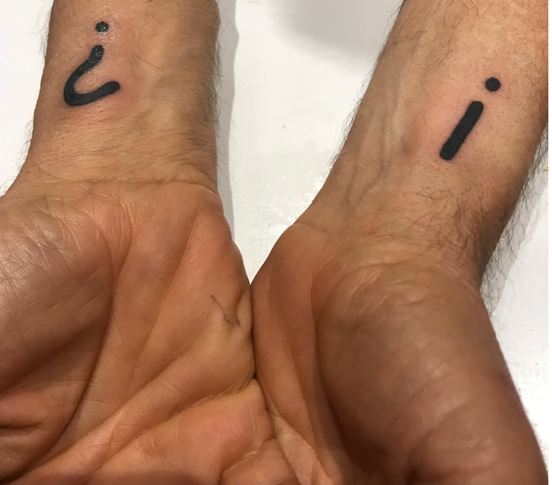 Two wrists side by side, each with a black tattoo: an inverted question mark on one and an inverted exclamation mark on the other